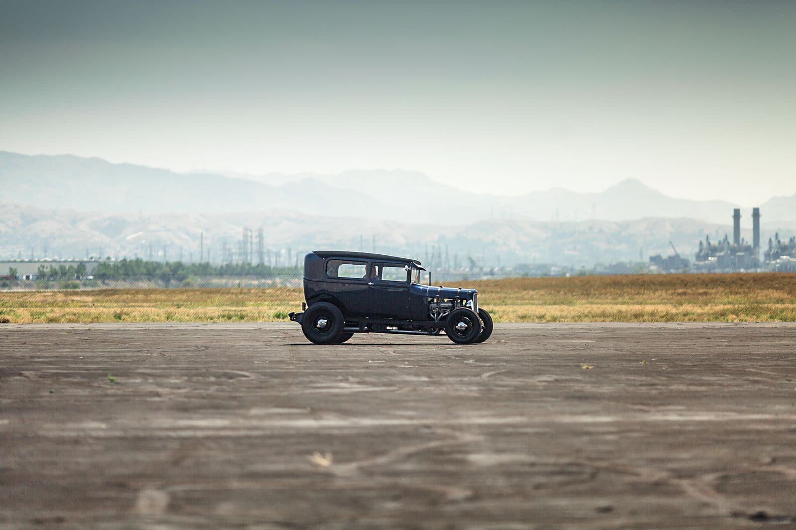 A chopped black hot rod sedan with an exposed engine parked on a wide concrete pad, with hazy hills behind it.