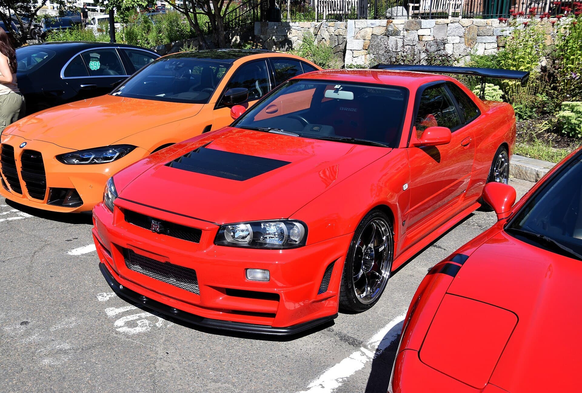 A red R34 Nissan Skyline GT-R with a black bonnet vent and a tall rear wing, parked beside other cars at an outdoor car meet.