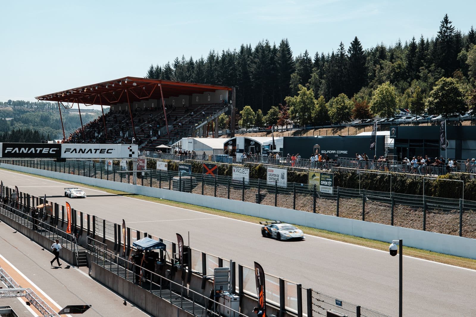 Two racing cars run down the main straight of a tree-lined circuit past a covered grandstand and pit buildings on a bright day.