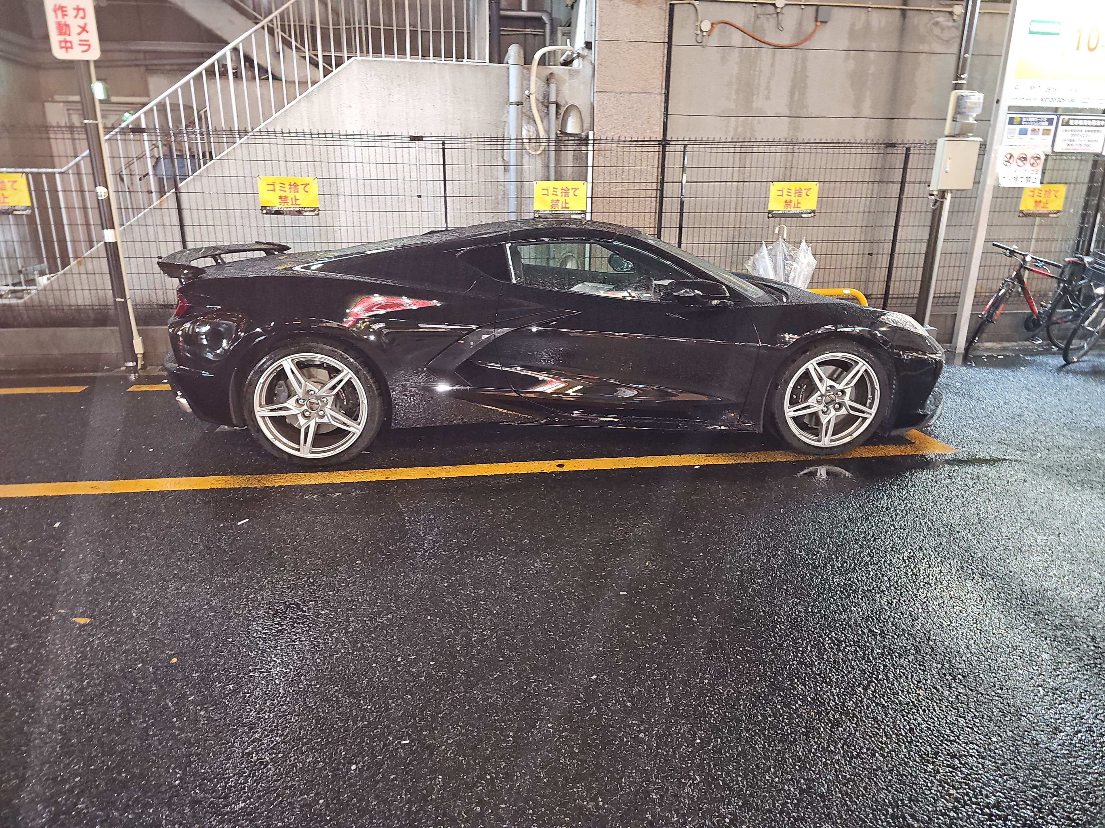 A black Chevrolet Corvette parked on wet pavement under lights in Osaka at night.