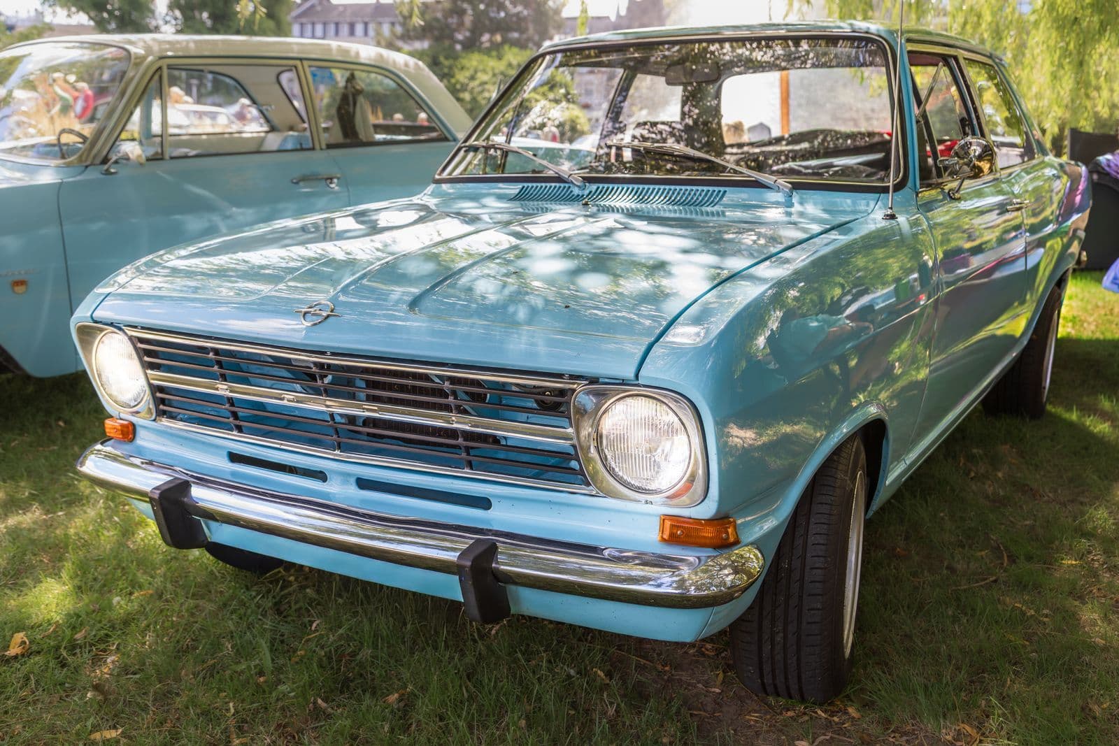 A pale blue vintage sedan displayed on grass under dappled shade at an outdoor car show.