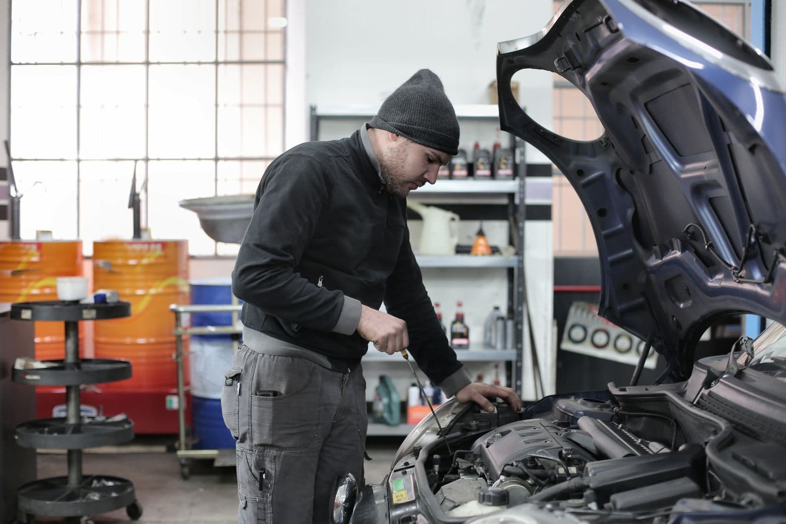 A man in a beanie leans over the open bonnet of a car in a workshop, checking the engine bay.