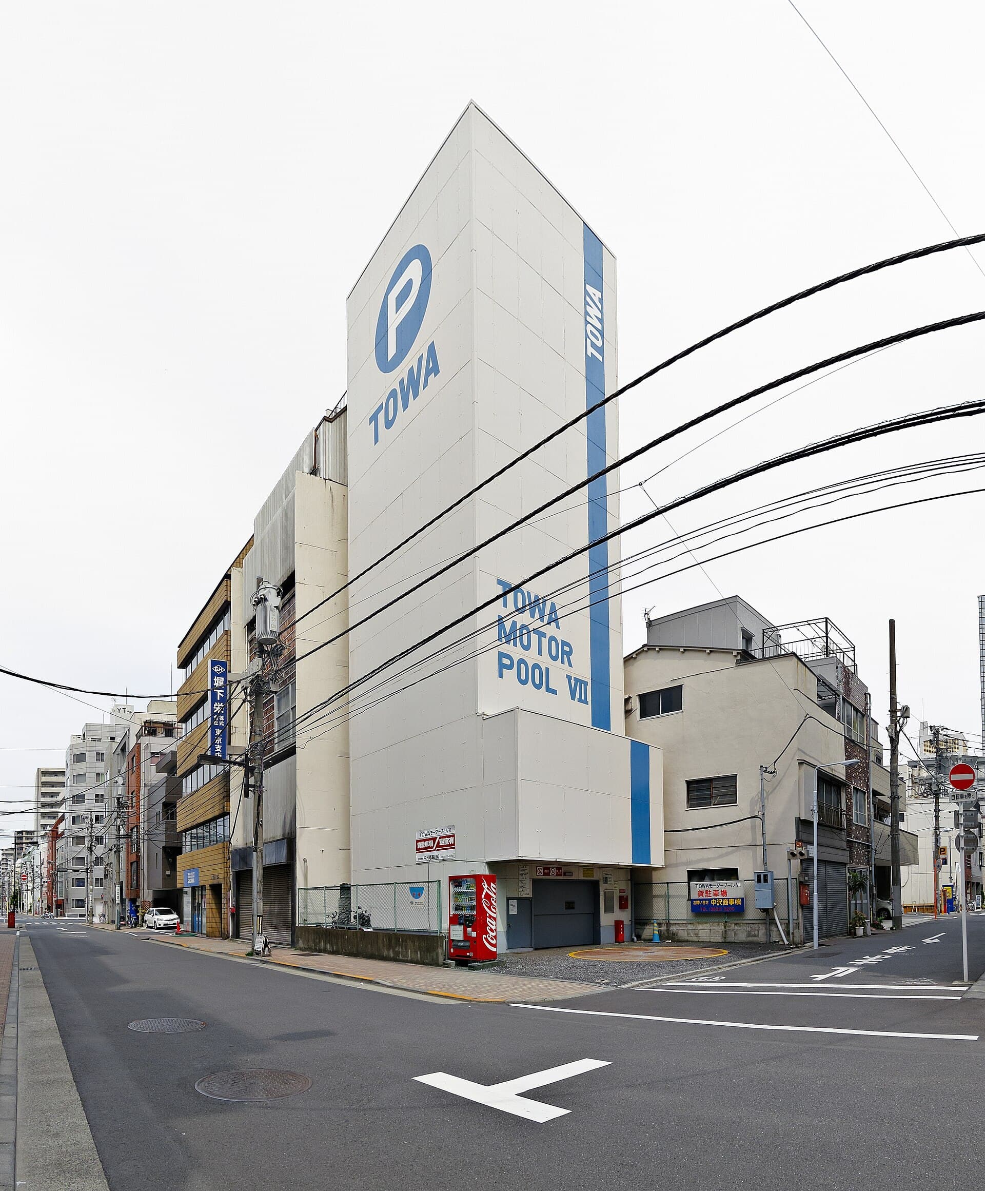 A tall, narrow white mechanical car-park tower branded TOWA Motor Pool VII, squeezed between low buildings on a Tokyo street corner, with a single vehicle door at its base.