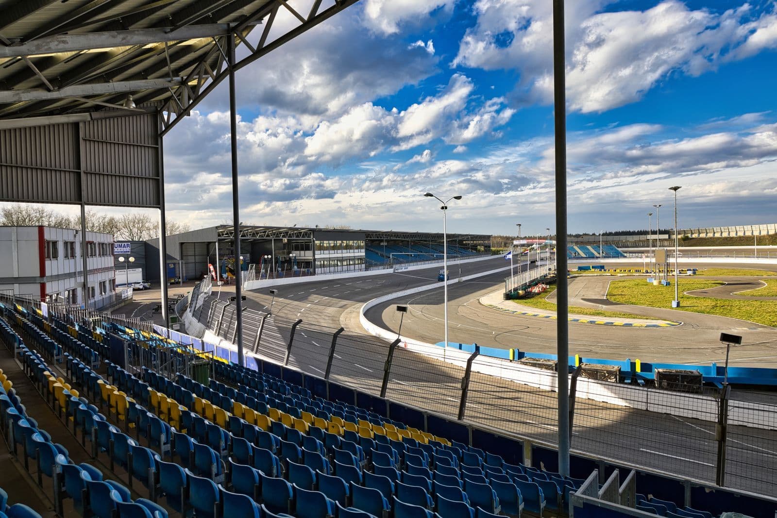 Empty blue and yellow grandstand seats overlooking a race circuit and pit buildings under a cloudy sky.