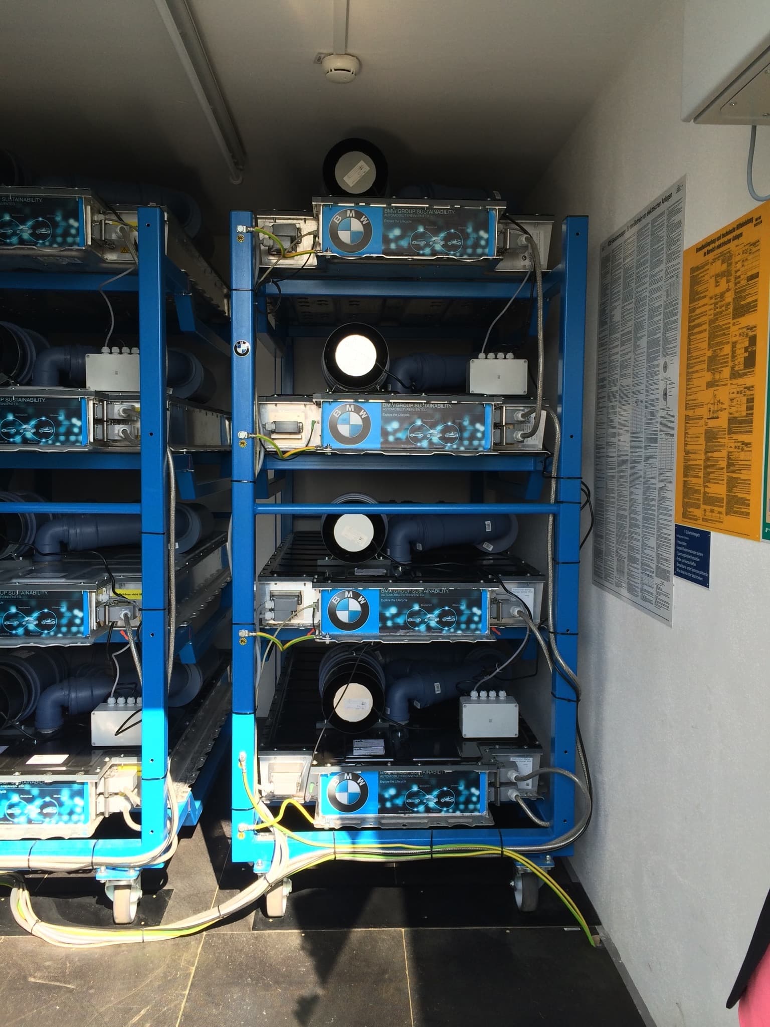 Blue metal racks holding stacked BMW-branded battery packs connected by cables in a small room.