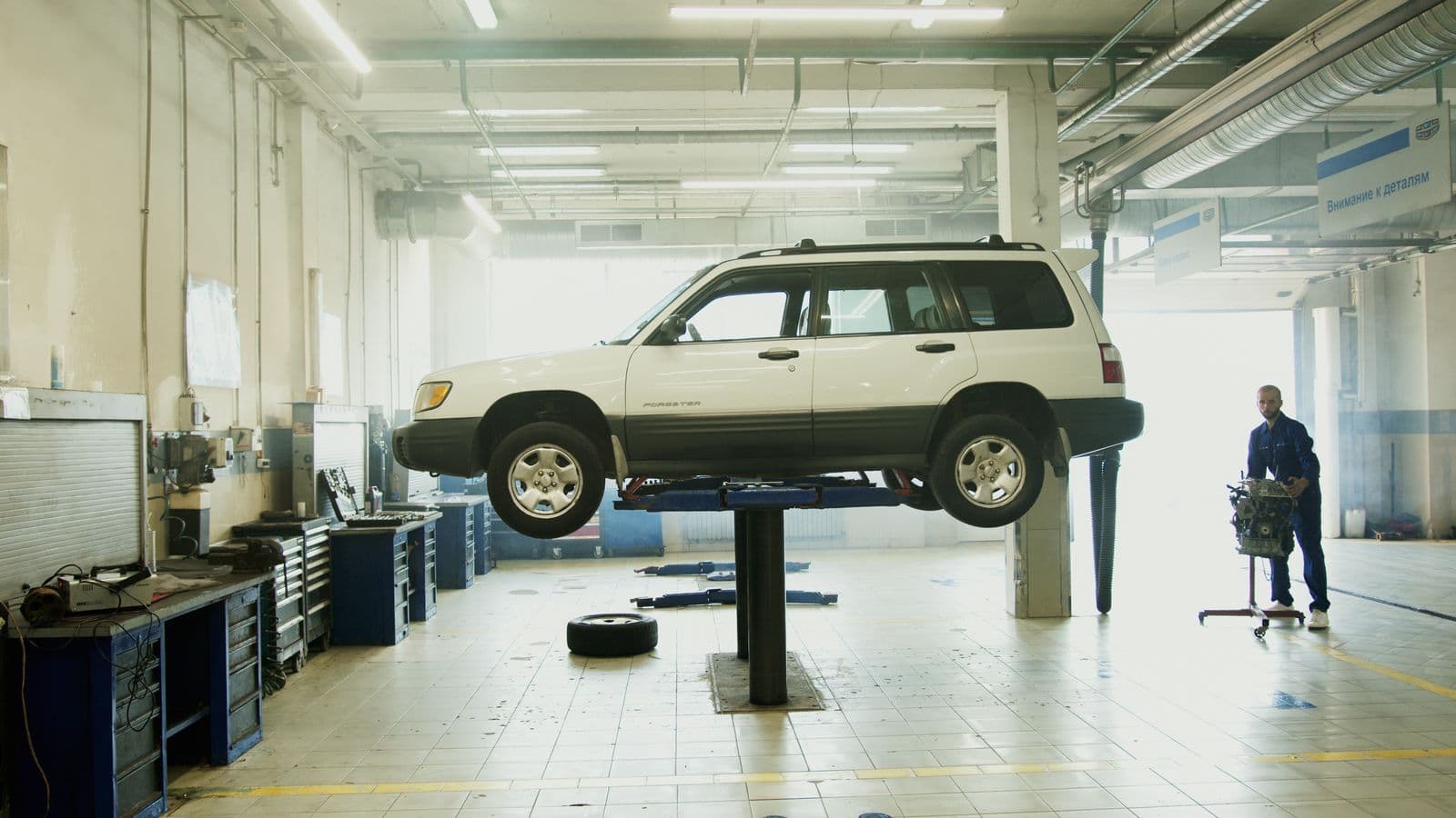 A white station wagon raised on a hoist in a workshop, with a wheel removed and a technician working nearby.
