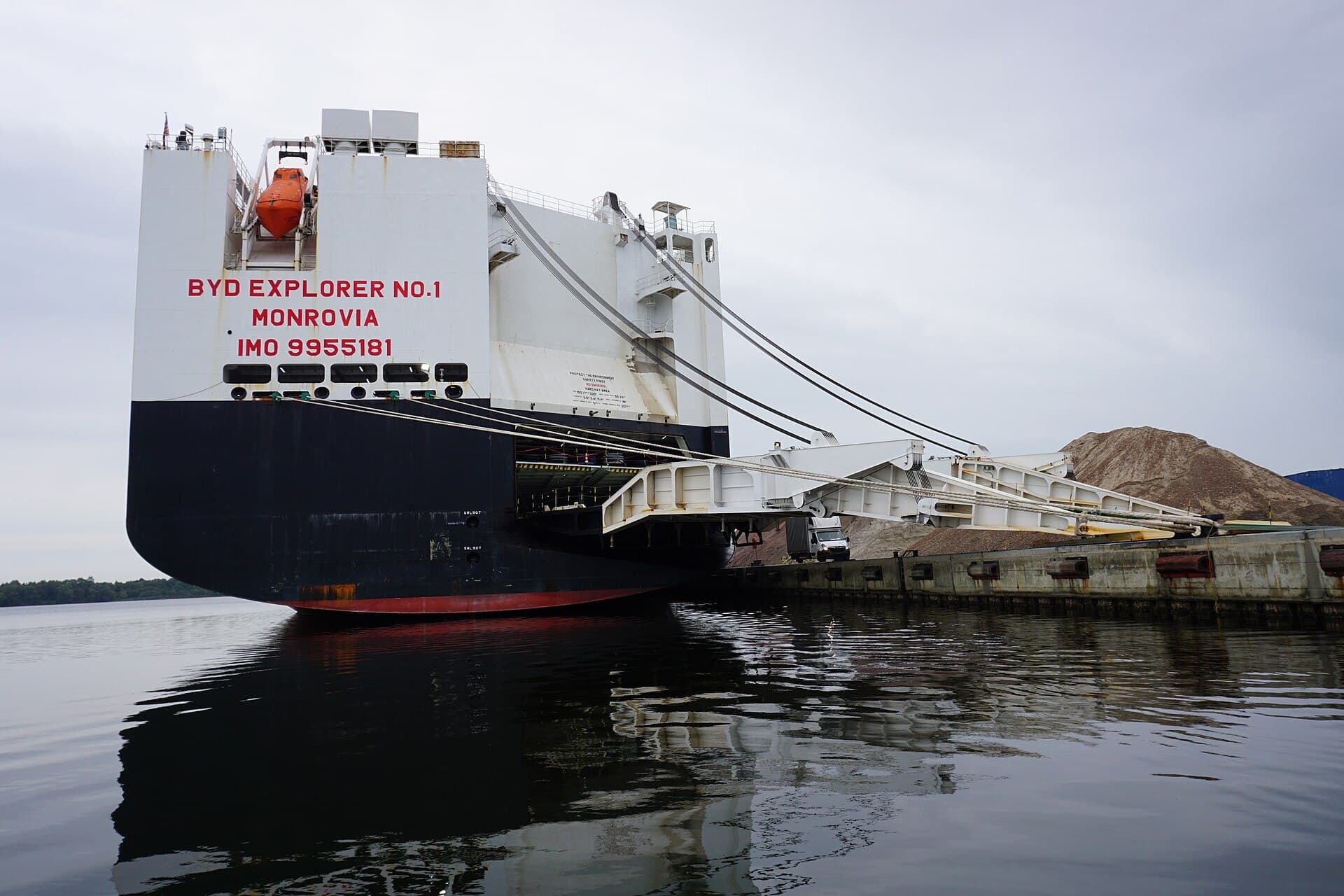 The white superstructure and dark hull of the car-carrier ship BYD Explorer No. 1, moored at a quay with its rear loading ramp lowered.