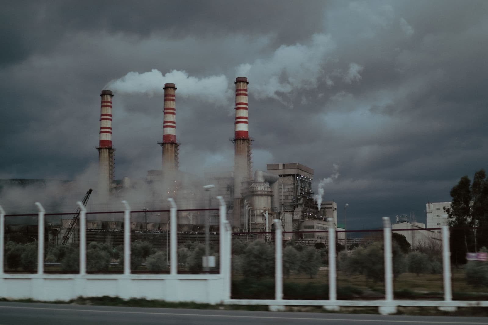 Three red-and-white striped chimneys of a power station releasing steam under dark storm clouds, seen from behind a roadside fence.