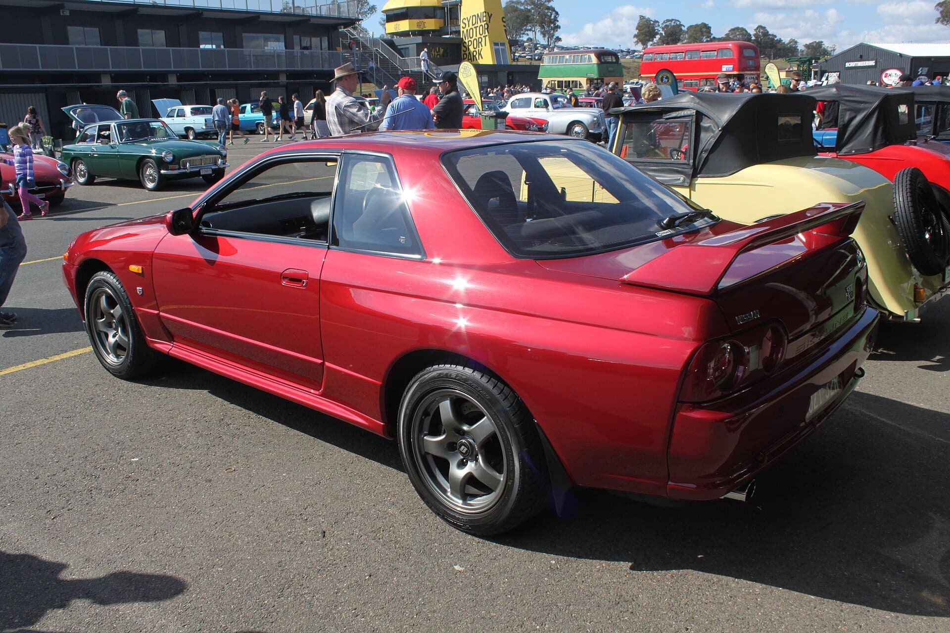 Rear three-quarter view of a red R32 Nissan Skyline GT-R coupe parked among classic cars at an outdoor show, with a grandstand and crowd in the background.