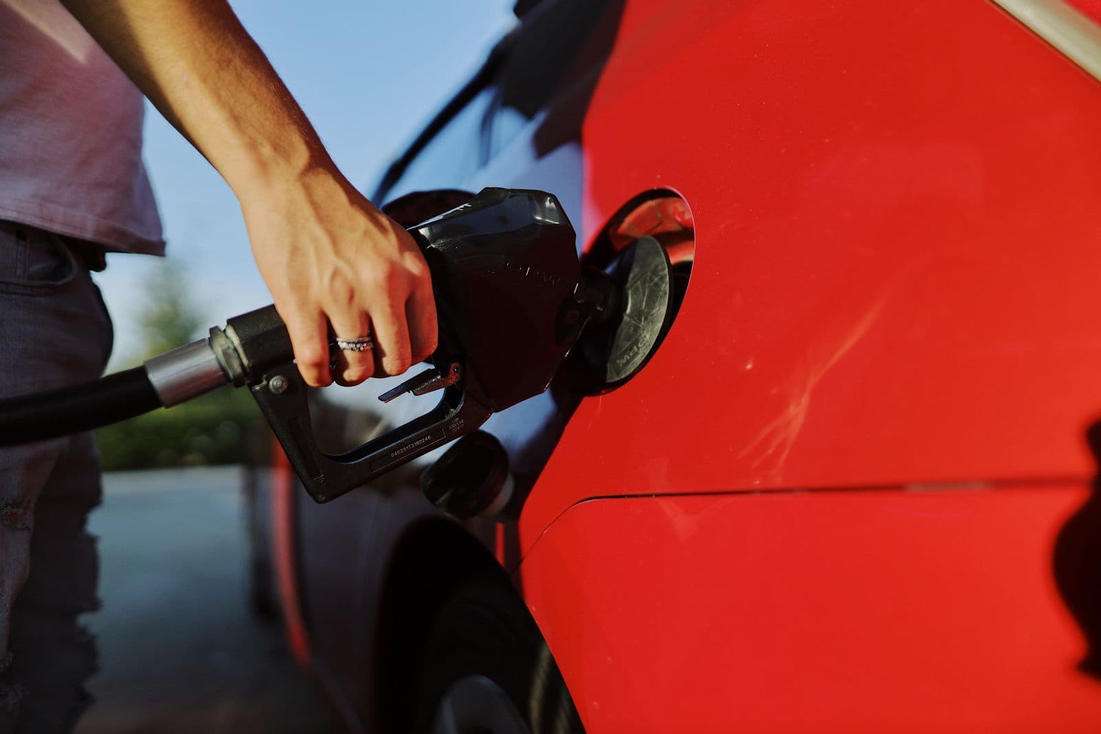 A hand holds a black fuel nozzle inserted into the open filler neck of a red car at a service station.