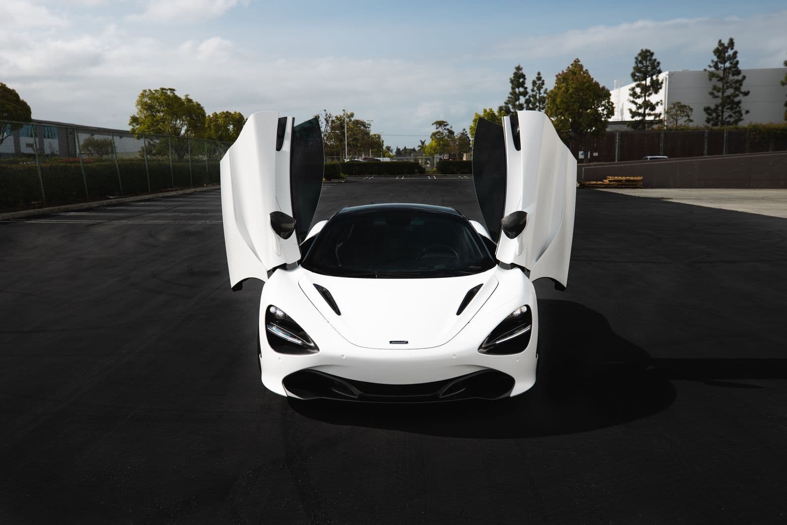 A white mid-engined supercar photographed head-on with both butterfly doors raised, standing on empty asphalt under a bright sky.
