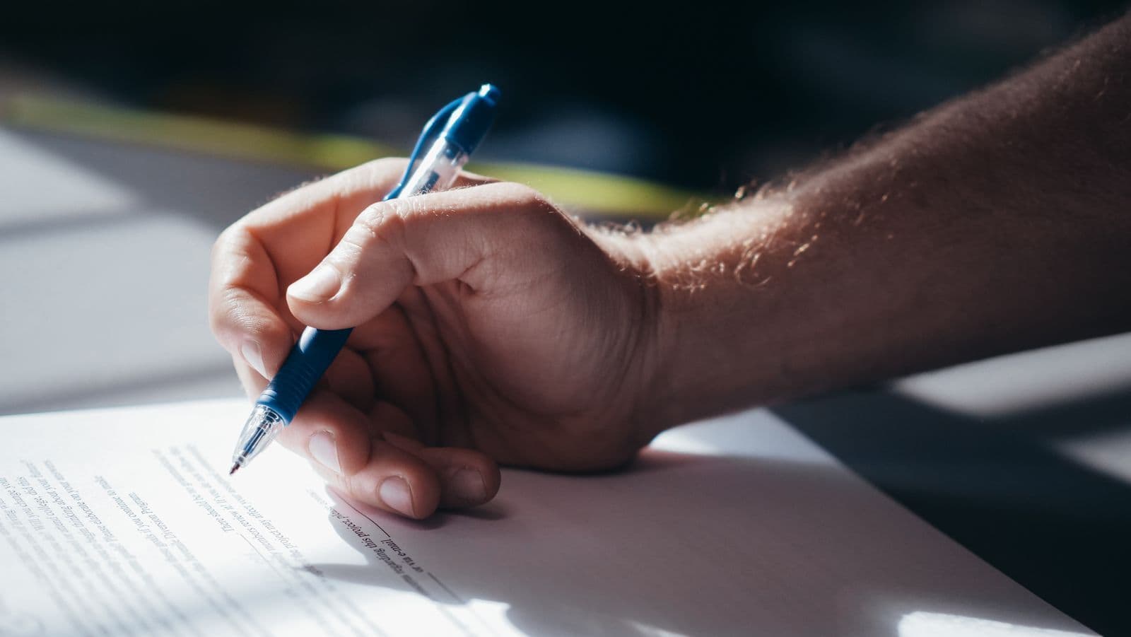 A hand holding a blue pen, signing a printed document on a desk in low, directional light.
