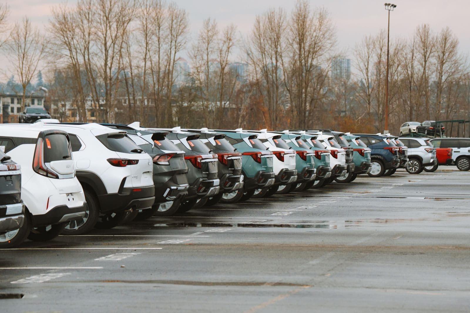 Long rows of new white, grey and teal SUVs parked tail-first in a wet dealership storage lot under an overcast sky.