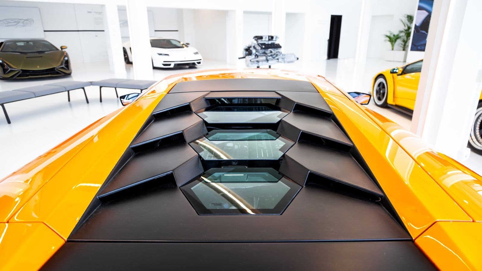The louvred engine cover of an orange mid-engined supercar in a white showroom, with other sports cars on display behind it.