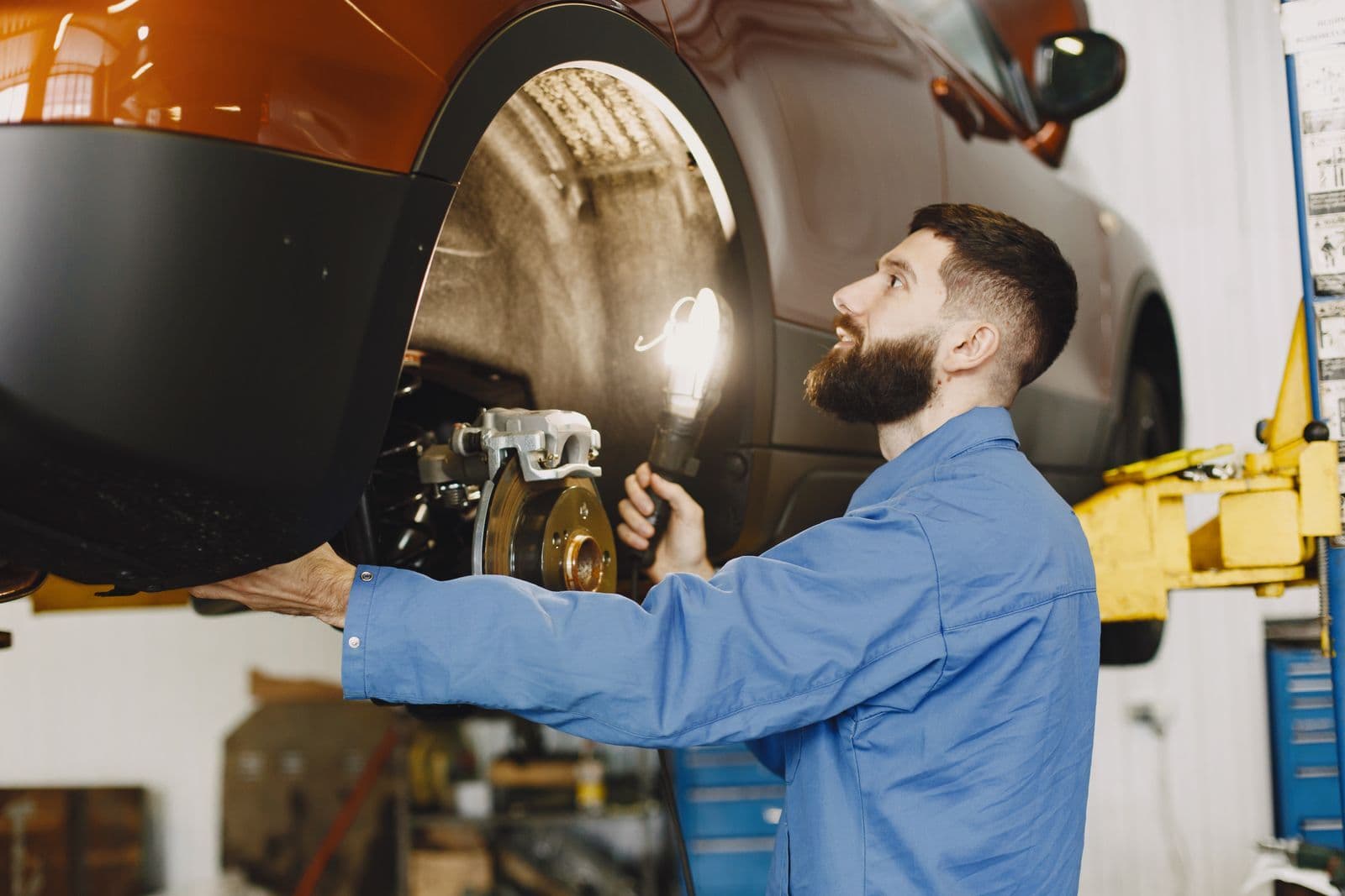 A mechanic in blue overalls shines a hand lamp on the brake disc of a car raised on a workshop hoist.