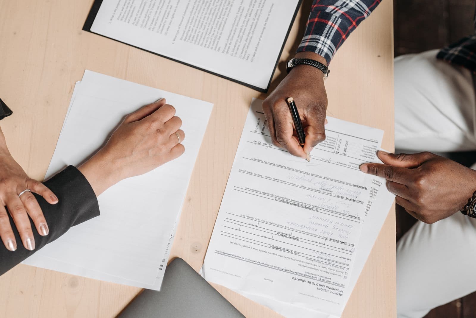 An overhead view of two people at a desk, one signing a printed form with a pen while the other rests a hand on a stack of papers.