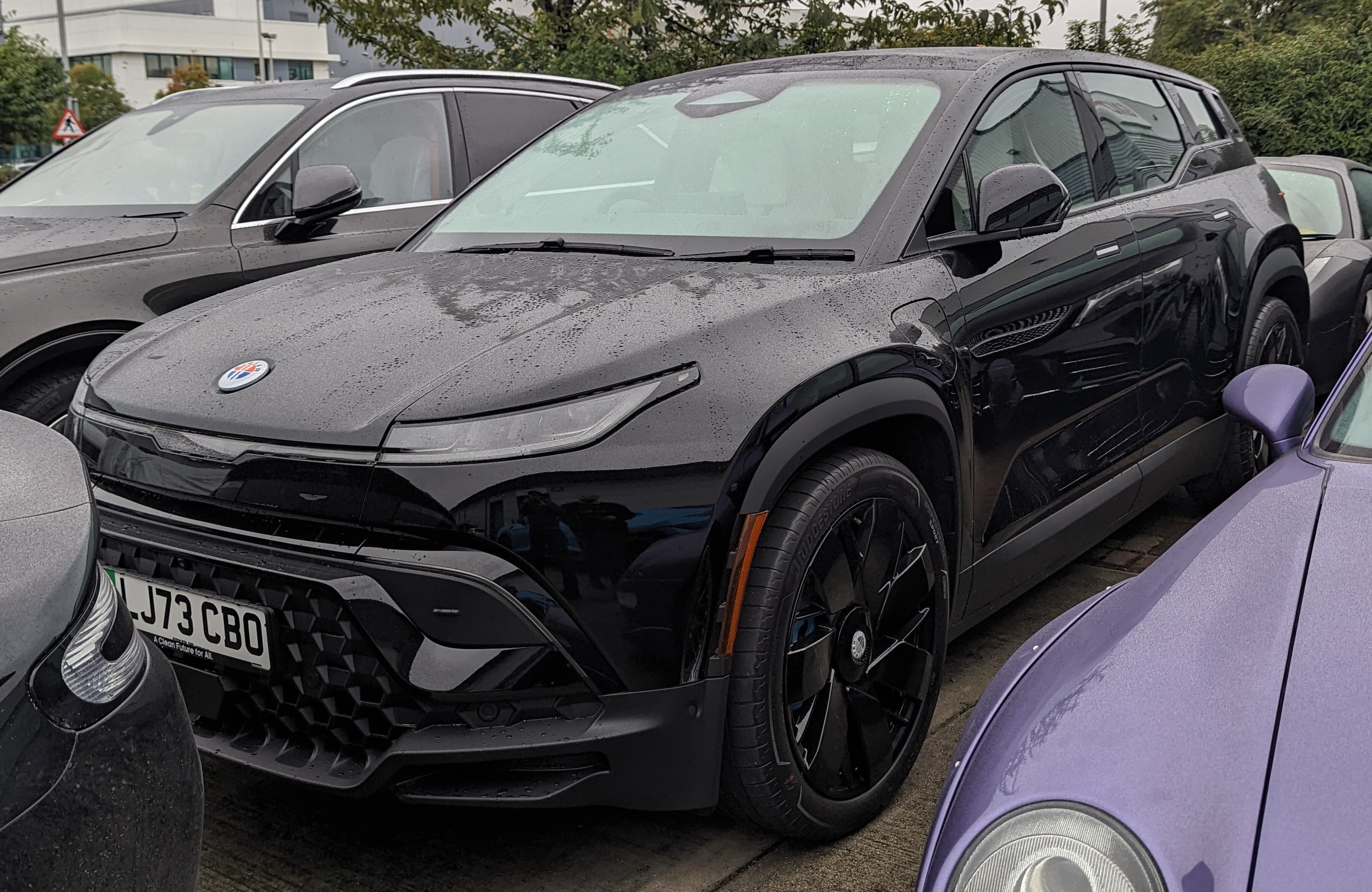 A dark Fisker Ocean SUV, covered in raindrops, parked closely between other cars.
