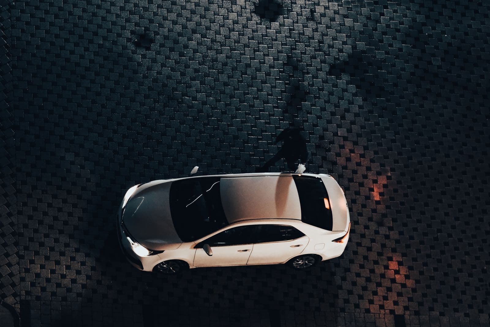 An overhead night view of a lone white hatchback parked on dark cobblestones, with a person's shadow falling beside it.