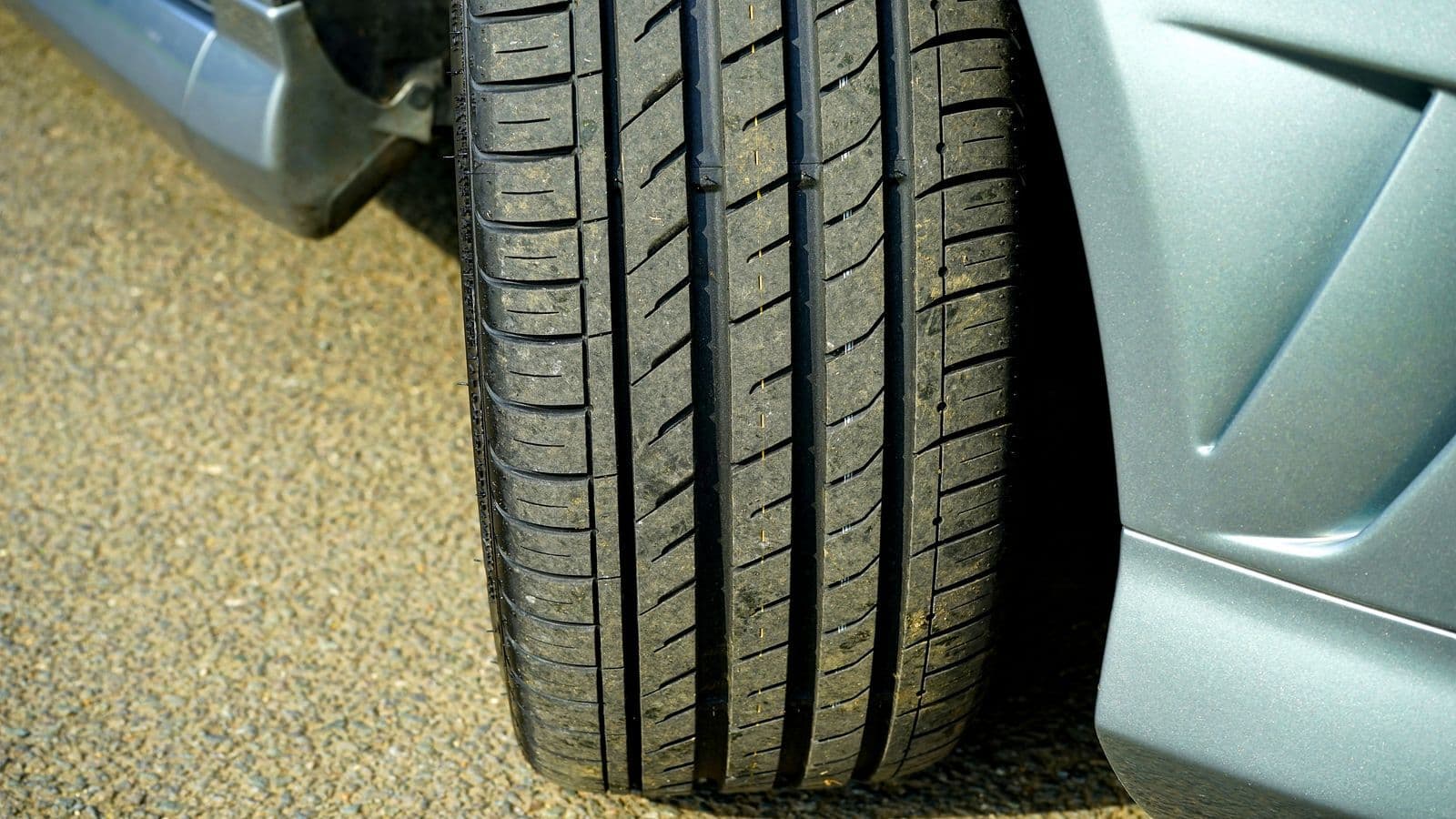 Close-up of a car tyre's tread pattern where it meets sunlit asphalt, beside a silver wheel arch.