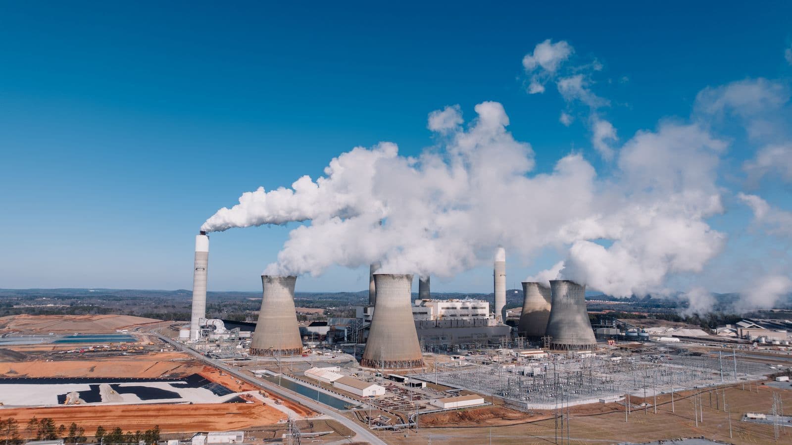 An aerial view of a coal-fired power station, its four cooling towers and tall stack venting white plumes into a clear blue sky above the switchyard.