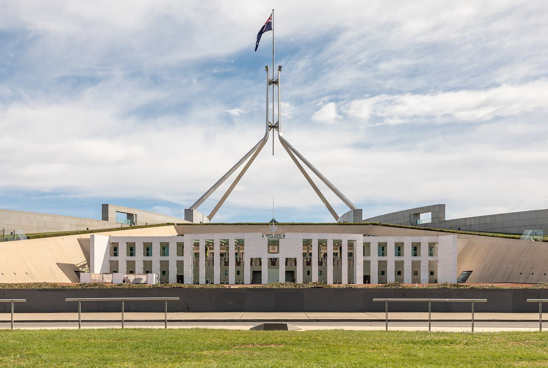 The front facade of Parliament House in Canberra under a cloudy blue sky, with the large four-legged flagpole flying the Australian flag.