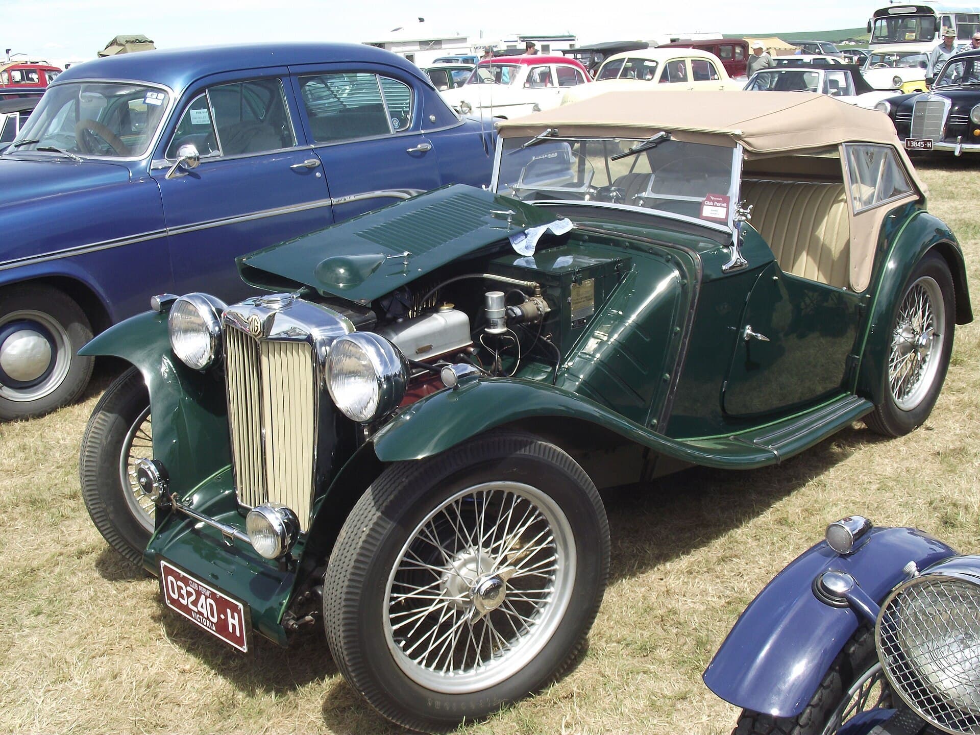 A green 1940 MG TB Midget with its bonnet open at an outdoor classic car display, with rows of other classic cars behind it