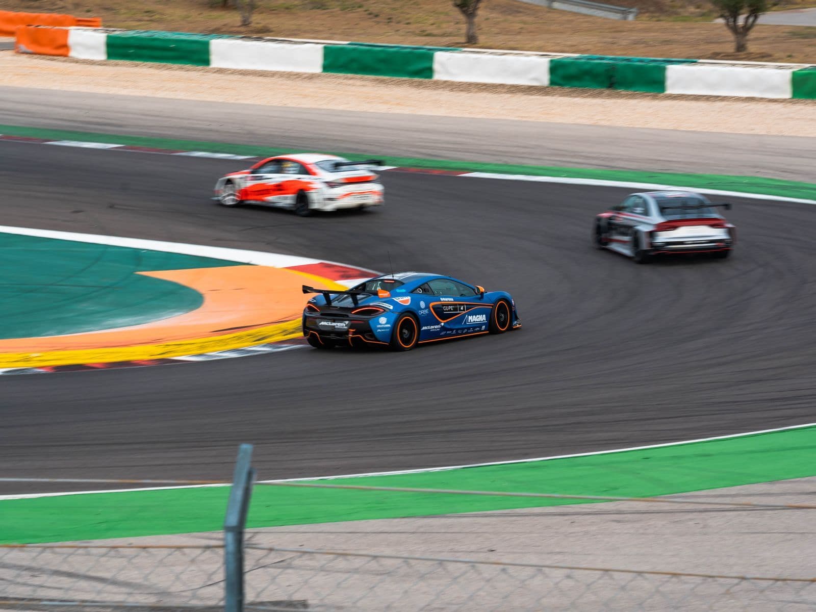 Three racing cars in team liveries sweep through a corner of a race circuit past coloured kerbs and trackside barriers.