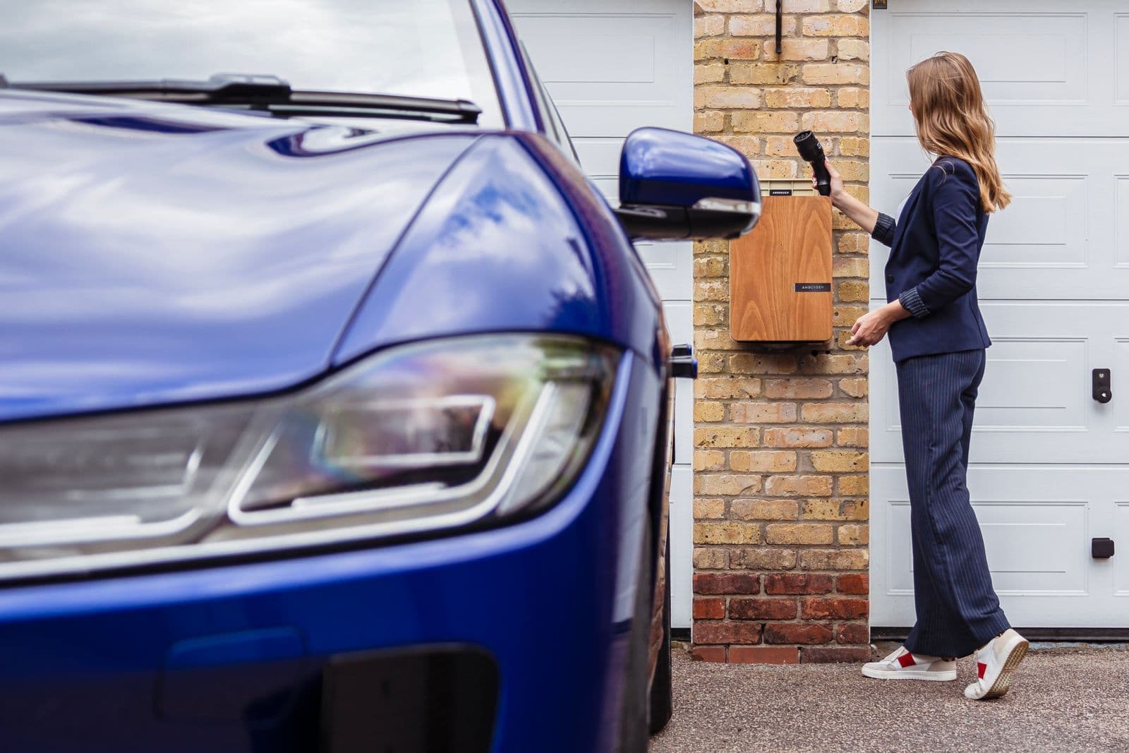 A woman lifts the cable from a wall-mounted home charger on a brick house wall, with a blue electric car parked on the driveway beside her.