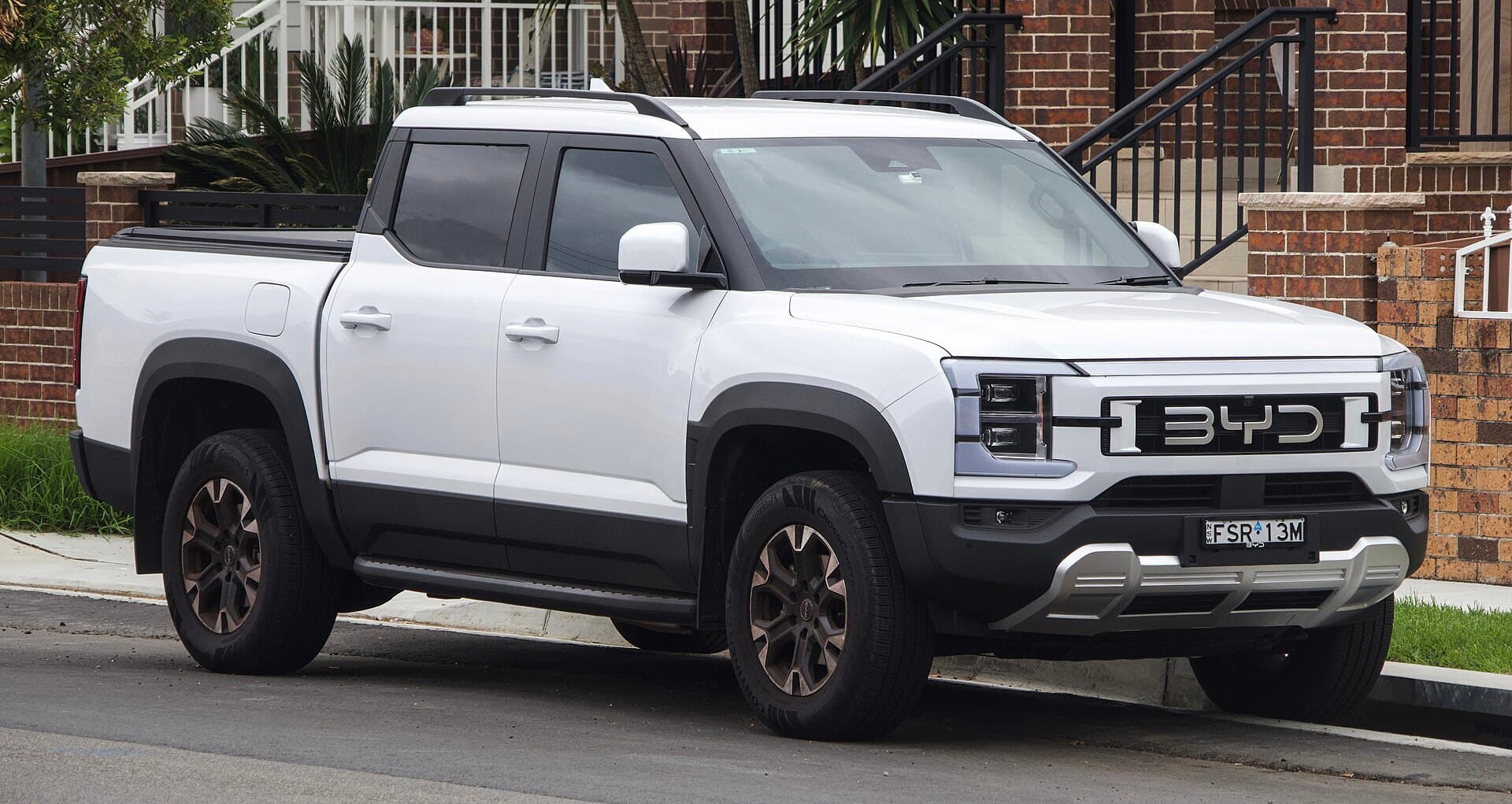 A white BYD Shark 6 dual-cab ute parked on a suburban street in front of brick houses.