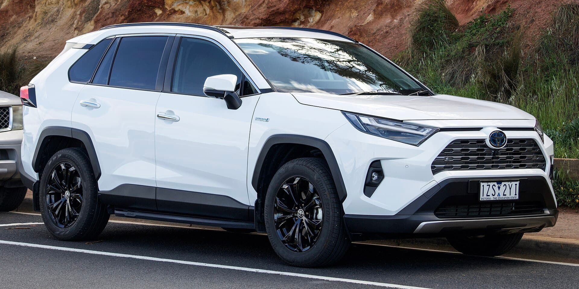 A white Toyota RAV4 Cruiser Hybrid SUV with black alloy wheels parked at a kerb beside a grassy cliff.