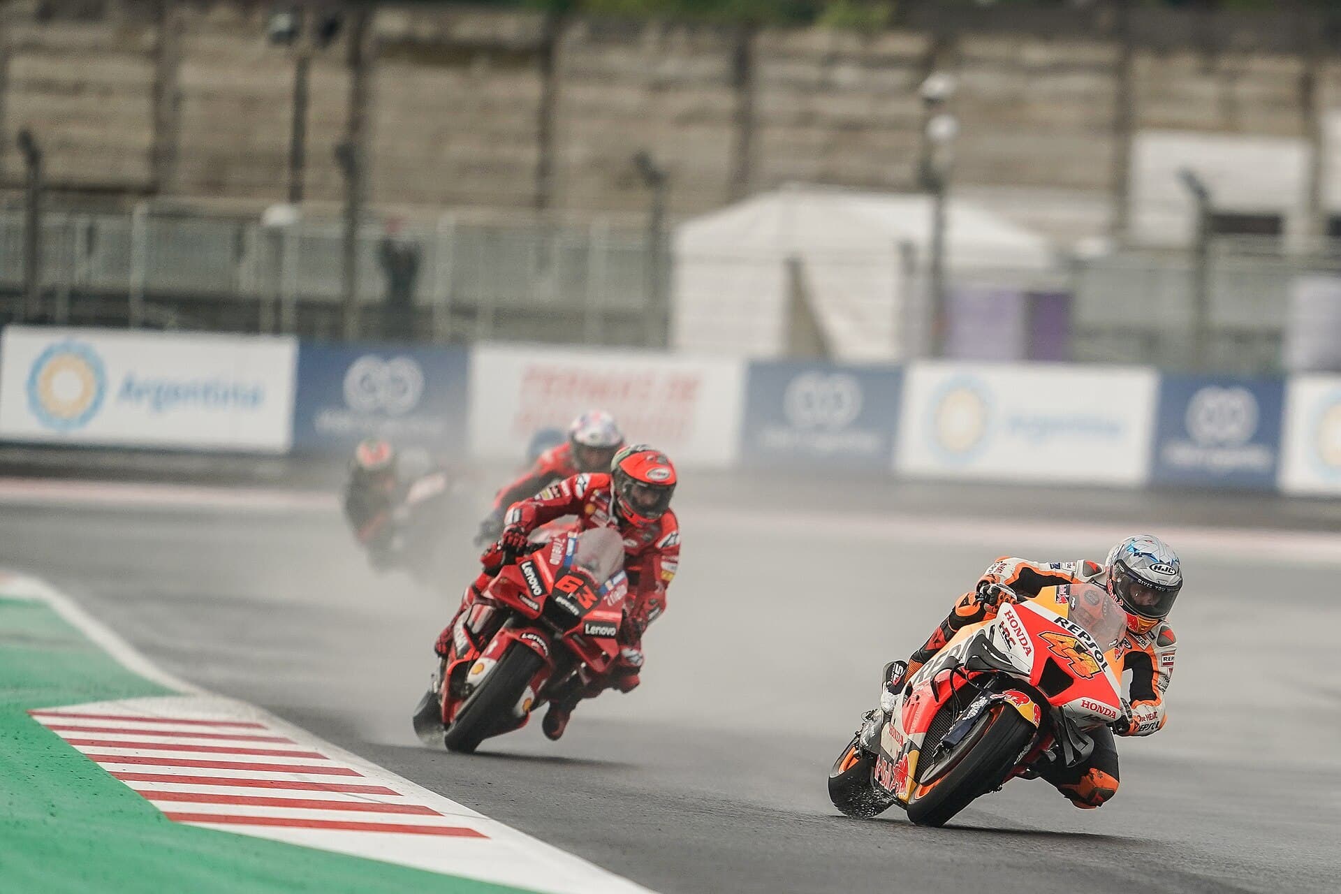 Four MotoGP riders lean their bikes through a corner on a wet, grey track, with spray behind them and empty grandstands in the background.