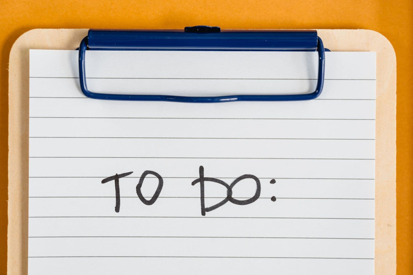 A wooden clipboard on an orange background holding a sheet of lined paper with the words "To Do:" written in black marker.
