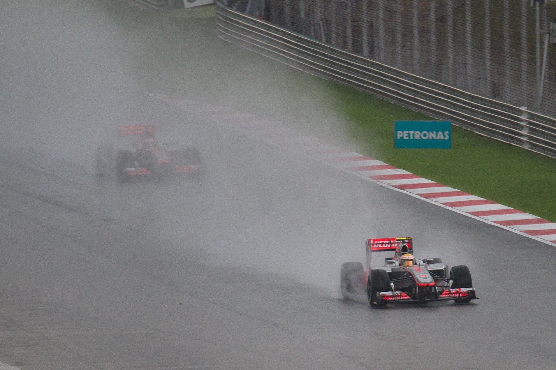 A silver and red Formula 1 car drives through heavy spray on a wet track, with a second car barely visible in the mist behind it.