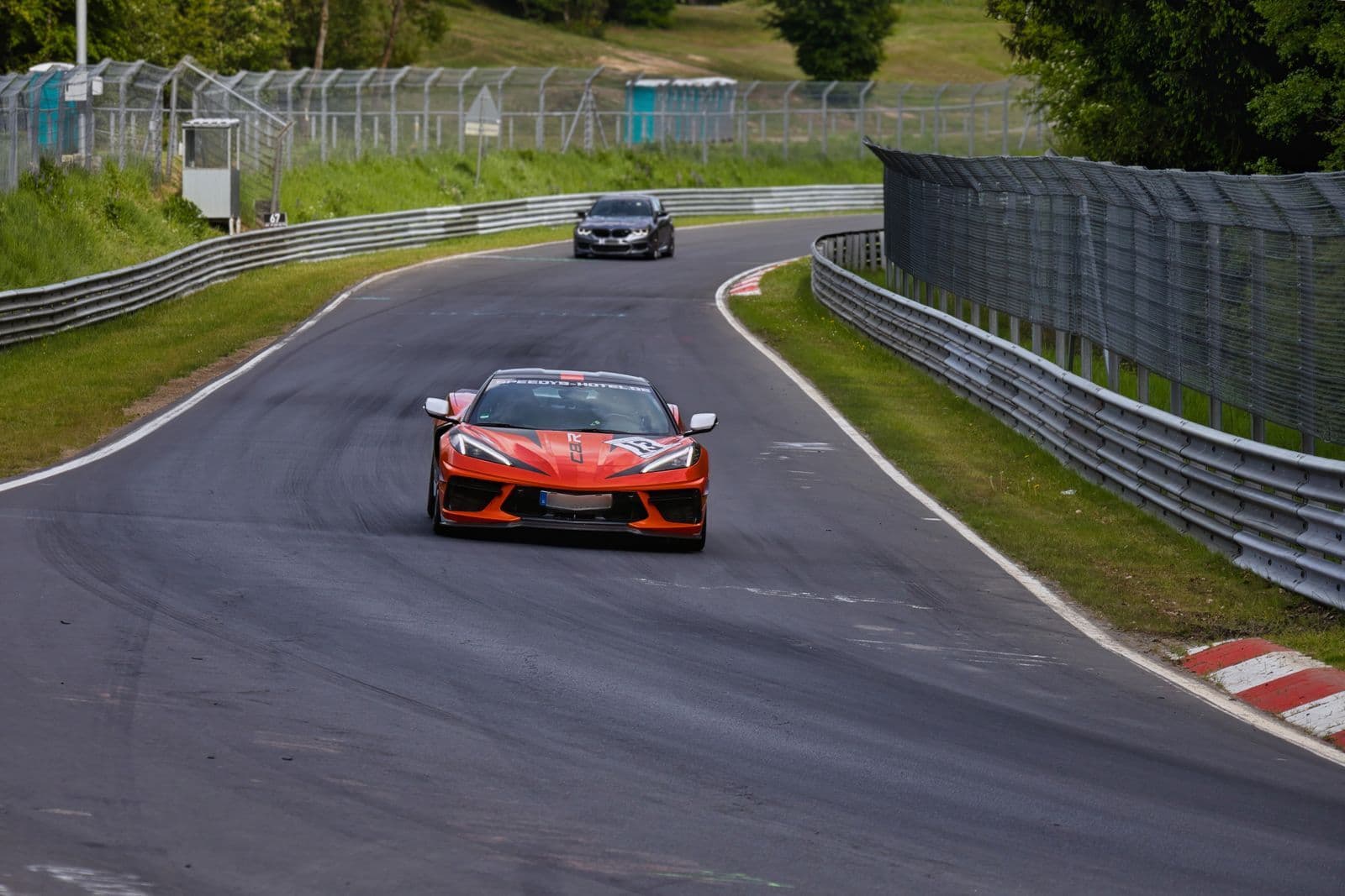 An orange sports car with racing decals corners on a circuit lined with steel barriers, a dark saloon following a few hundred metres behind.