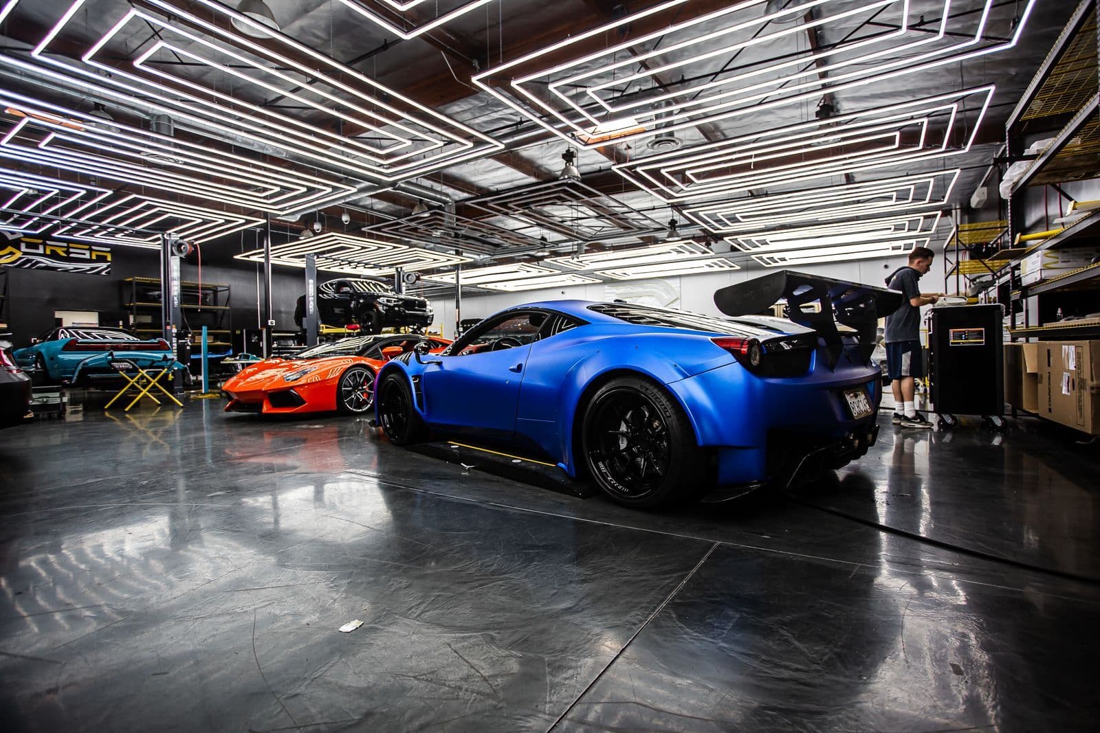 A matte blue supercar parked in a brightly lit specialist workshop alongside an orange supercar and other exotic cars.