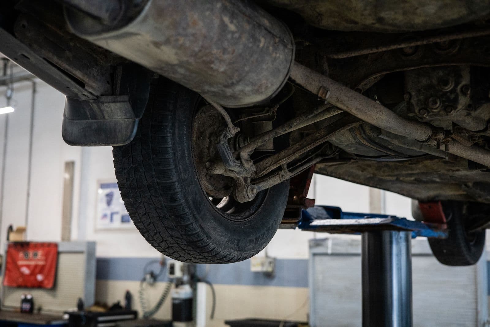 The underside of a car raised on a workshop hoist, showing a rear tyre, suspension arms and the exhaust.
